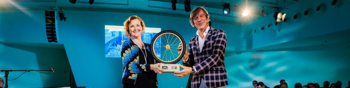 Martina Huijsmans, portfeuillehouder Duurzame Mobiliteit bij de MRDH ontvangt van directeur CROW, Pieter Litjens het gouden wiel. De prijs. Ze staan op een podium en houden de prijs beiden vast.
