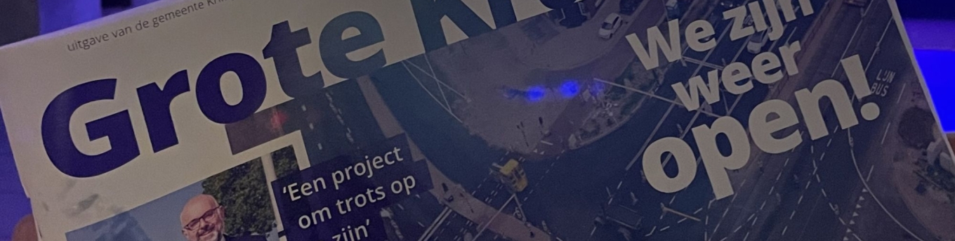 Iemand laat het magazine van de opening van de Grote Kruising zien. Op de achtergrond speelt een band op het podium tijdens de opening van de Grote Kruising