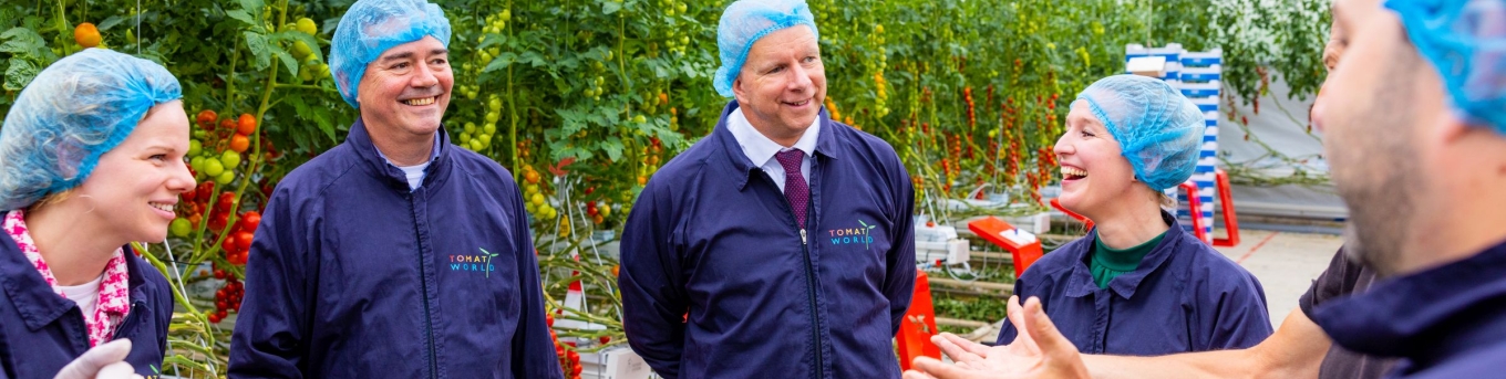 Gedeputeerde Meindert Stolk, wethouder Pieter Varekamp (Westland) en wethouder Maaike Zwart (Delft) zijn in gesprek met medewerkers van TomatoWorld. Ze staan in een kas met beschermende kleding aan.