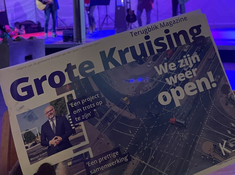 Iemand laat het magazine van de opening van de Grote Kruising zien. Op de achtergrond speelt een band op het podium tijdens de opening van de Grote Kruising