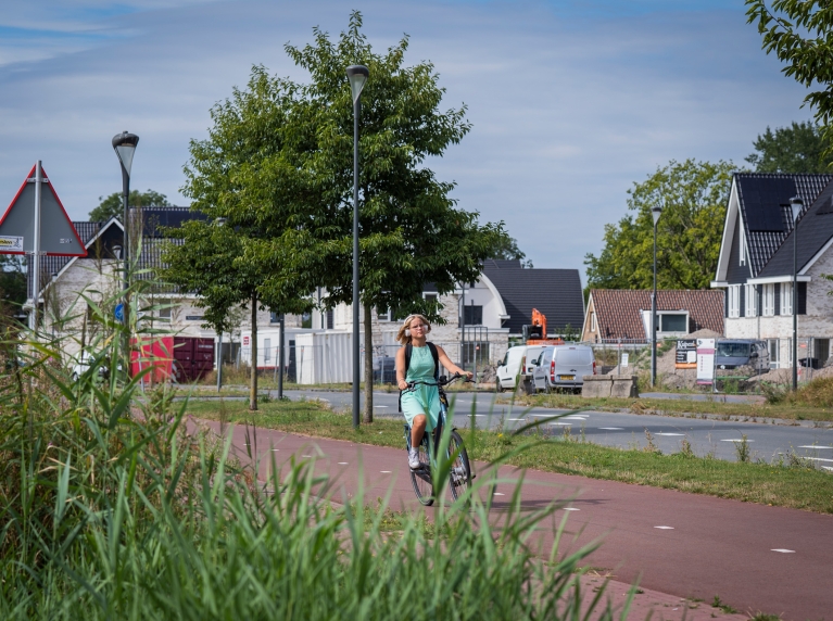 Fietsster fietst op een vrijliggend geasfalteerd roodkleurig fietspad. Op de achtergrond zijn woningen te zien.